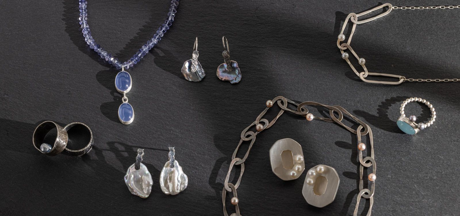 Danni’s jewelry laid out on dark slate: a blue chalcedony and iolite necklace, pearl earrings, two rings, silver link necklaces and pearl studs